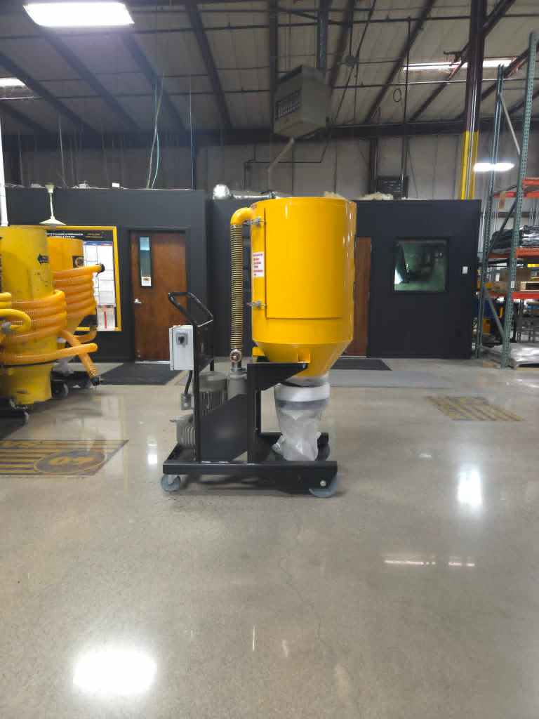 Yellow industrial machine on a wheeled stand in a factory, with orange hoses and dark partition walls in the background.