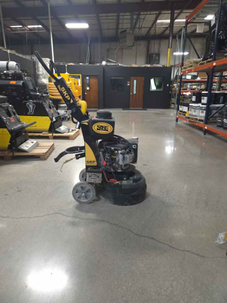 Yellow floor-grinding machine in a concrete warehouse, with shelving and dark partitioned offices in the background.