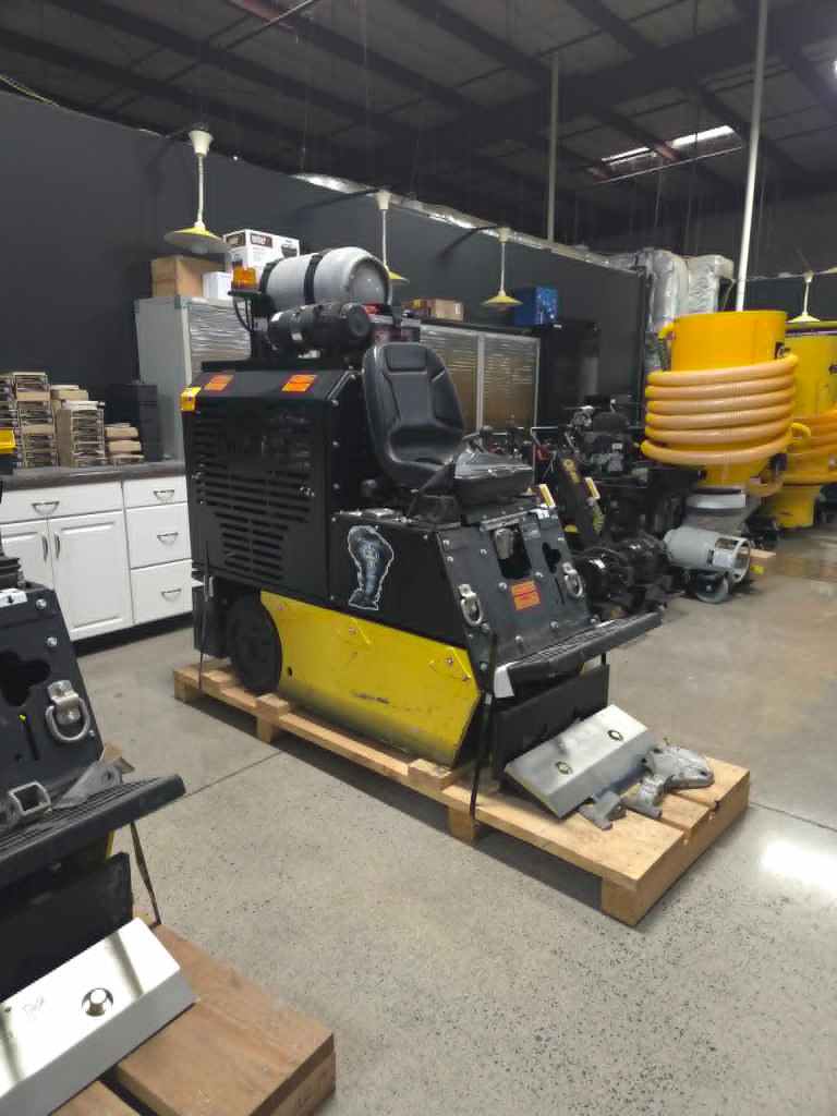 Industrial workshop scene with a large yellow and black machine on a wooden pallet, coiled orange hose nearby, and shelves in the background.
